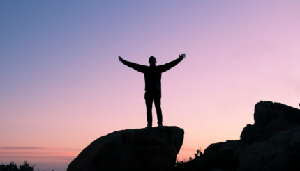 man stood on large rock with arms up in celebration at daybreak