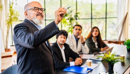 Smart Suit Wearing Office Director Holding A Pen and Presenting To Group of Younger Colleagues
